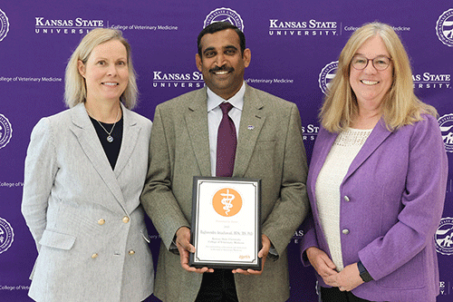 Three individuals stand proudly in front of a purple banner displaying an award, smiling at the camera.