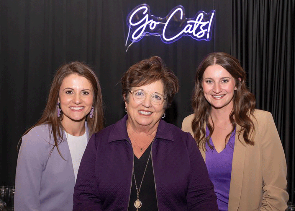Three women standing together, smiling, at a social event, dressed in formal attire.