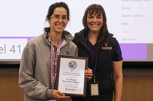 Two women celebrate with an award, positioned in front of a screen that features visual information.