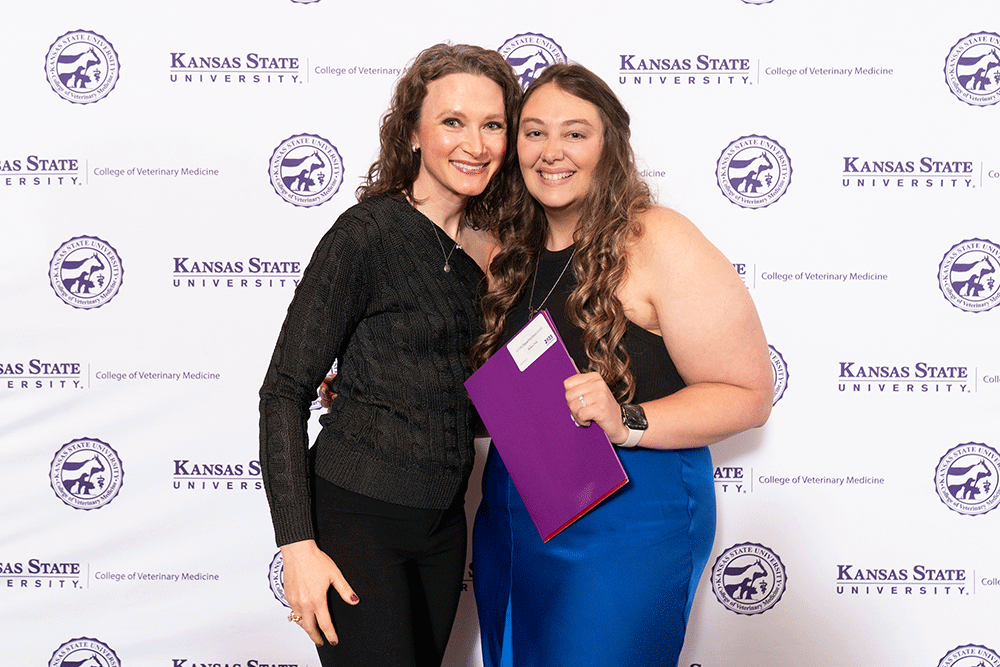 Two women stand in front of a backdrop smiling.