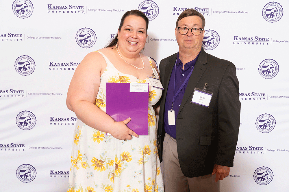 Two people stand in front of a backdrop smiling.