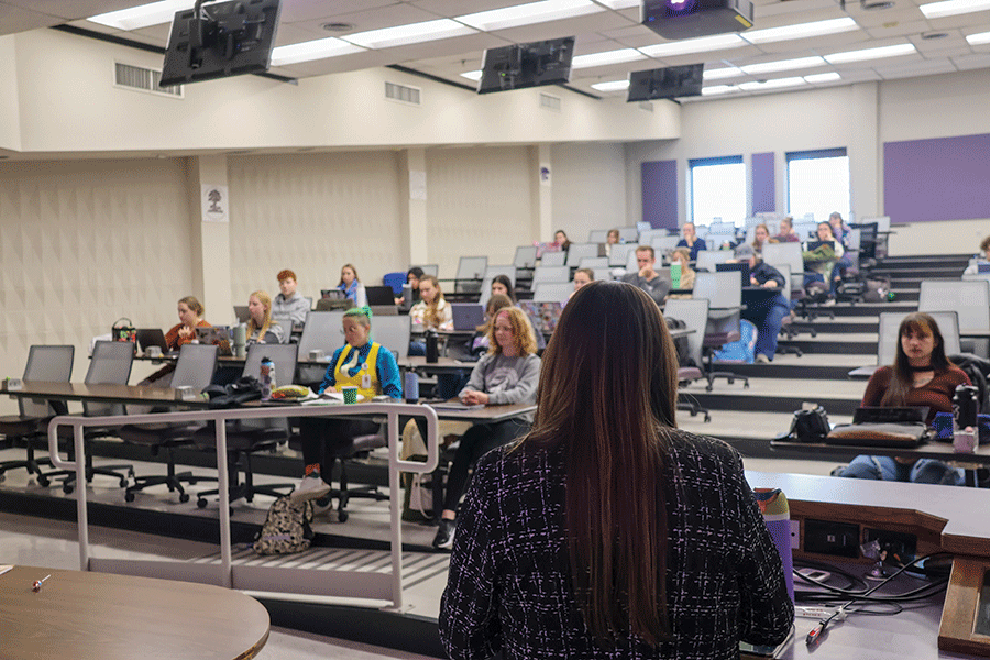 A woman lectures in a classroom, engaging students who are seated and focused on her presentation.