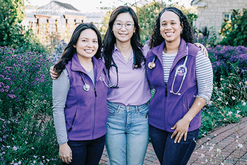 Three women wearing purple vests with stethoscopes around their necks stand together smiling in a garden.