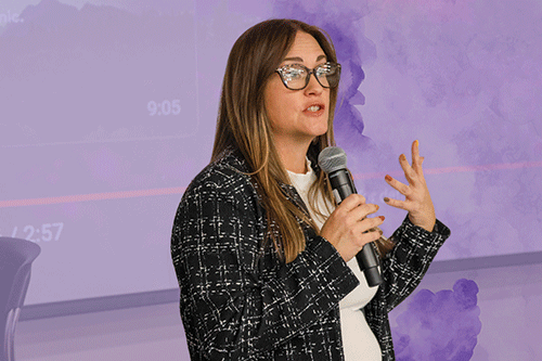 A woman wearing glasses stands confidently while giving a presentation to an audience.