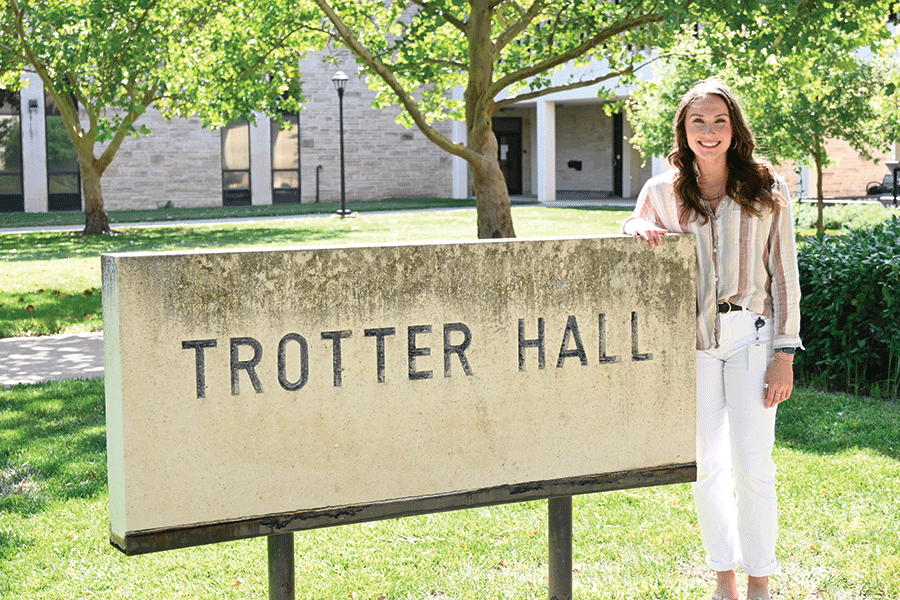 A woman stands beside a sign reading "Trotter Hall," smiling and dressed casually in a sunny outdoor setting.