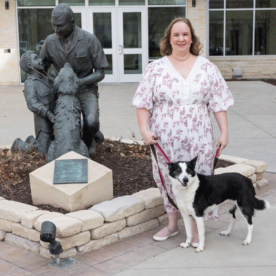 A woman stands beside her dog, both facing a prominent statue in an outdoor area.