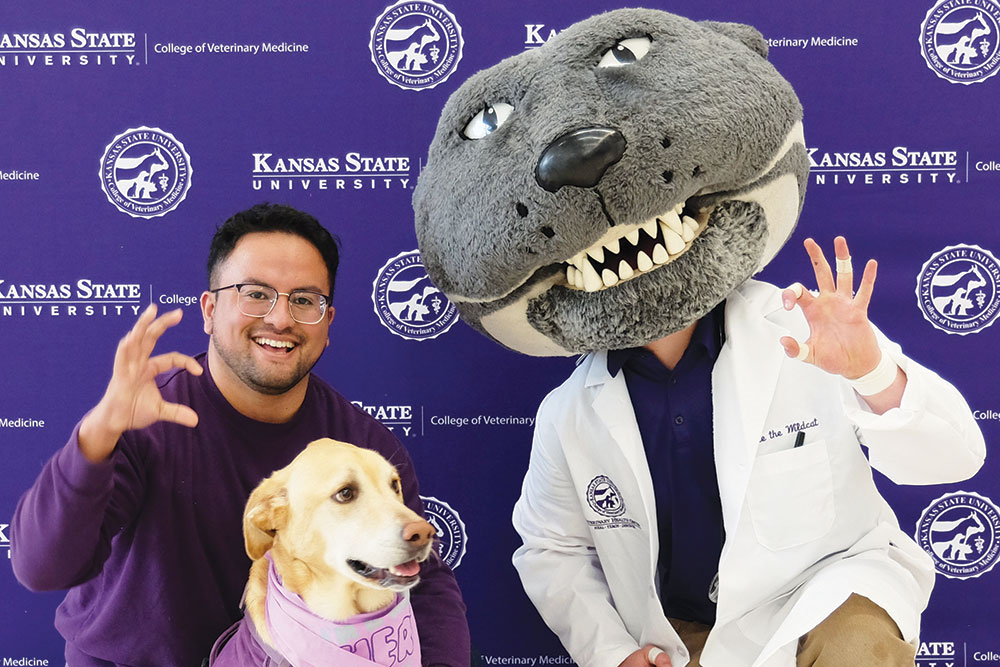 Willie the Wildcat poses with a man and his dog in front of a purple backdrop.